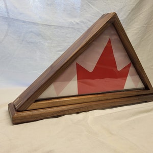Peut inclure: Un présentoir de drapeau en bois de forme triangulaire. Le boîtier est en bois foncé et possède une façade en verre. À l'intérieur se trouve un drapeau canadien avec une feuille d'érable rouge sur fond blanc.