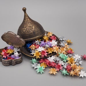 Könnte beinhalten: Eine antike Messingdose mit Deckel und aufwendigen Blumendekoren, geöffnet, um eine Sammlung bunter Origami-Sterne zu enthüllen.