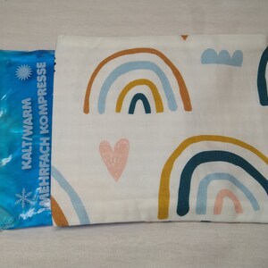 Könnte beinhalten: Ein weißer Stoffbeutel mit einem blauen Gelpack im Inneren. Der Beutel hat ein Regenbogenmuster mit Herzen und den Text "KALT/WARM MEHRFACH KOMPRESSE".