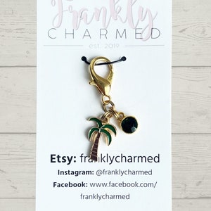 May include: A gold palm tree charm with a black gem, attached to a gold lobster clasp and black string. The charm is on a white card with the text "Frankly CHARMED est. 2019" and social media information for the shop.
