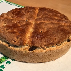 May include: A round loaf of Irish soda bread, golden brown with a cross cut into the top. The bread has a slightly rough texture and visible raisins. It sits on a white cloth with a green shamrock border, suggesting a St. Patrick's Day theme.