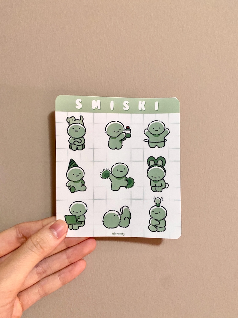 May include: A square sticker sheet featuring nine green cartoon figures in various poses. The word "SMISKI" is printed at the top. The figures are on a white grid background.