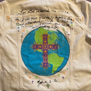 Könnte beinhalten: Eine weiße Jeansjacke mit einem gemalten Bild der Erde und einem Kreuz. Der Text "For God so loved the world, that He gave His only begotten Son that whosoever believeth in Him should not perish, but have everlasting life. John 3:16" ist in schwarzer Farbe auf der Rückseite der Jacke geschrieben.