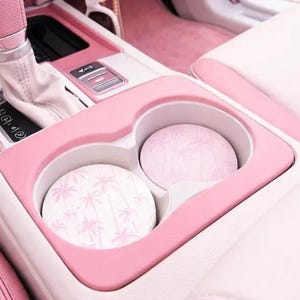 May include: A car interior featuring a pink center console with two circular cup holder inserts. The inserts are white with a repeating light pink palm tree pattern. A pink gear shift knob and white sunglasses are visible near the dashboard area.
