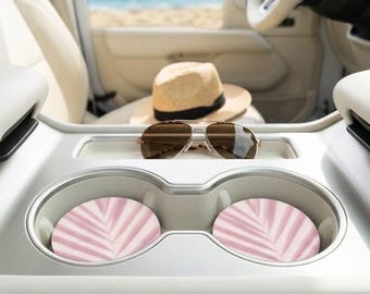 Può includere: Interno auto con cappello di paglia, occhiali da sole e due sottobicchieri rosa e bianchi con motivo a foglie. Lo sfondo mostra una spiaggia e l'oceano, suggerendo un tema estivo.