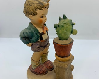 Może przedstawiać: Porcelanowa figurka chłopca w zielono-czarnej kratkowanej koszuli i brązowych spodenkach, patrzącego na zielonego kaktusa w brązowej doniczce. Figurka stoi.