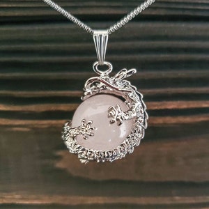 Pode incluir: Um colar com um pingente de dragão prateado e uma esfera de quartzo rosa. O dragão está enrolado em torno da esfera, com a cauda envolvendo a parte frontal.
