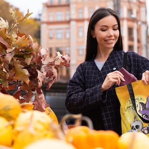 May include: A woman is holding a yellow tote bag with a black cat wearing a witch hat and the text "It's a Witch Thing". The bag is in front of a pile of pumpkins.