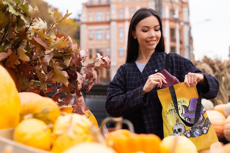 May include: A woman is holding a yellow tote bag with a black cat wearing a witch hat and the text "It's a Witch Thing". The bag is in front of a pile of pumpkins.