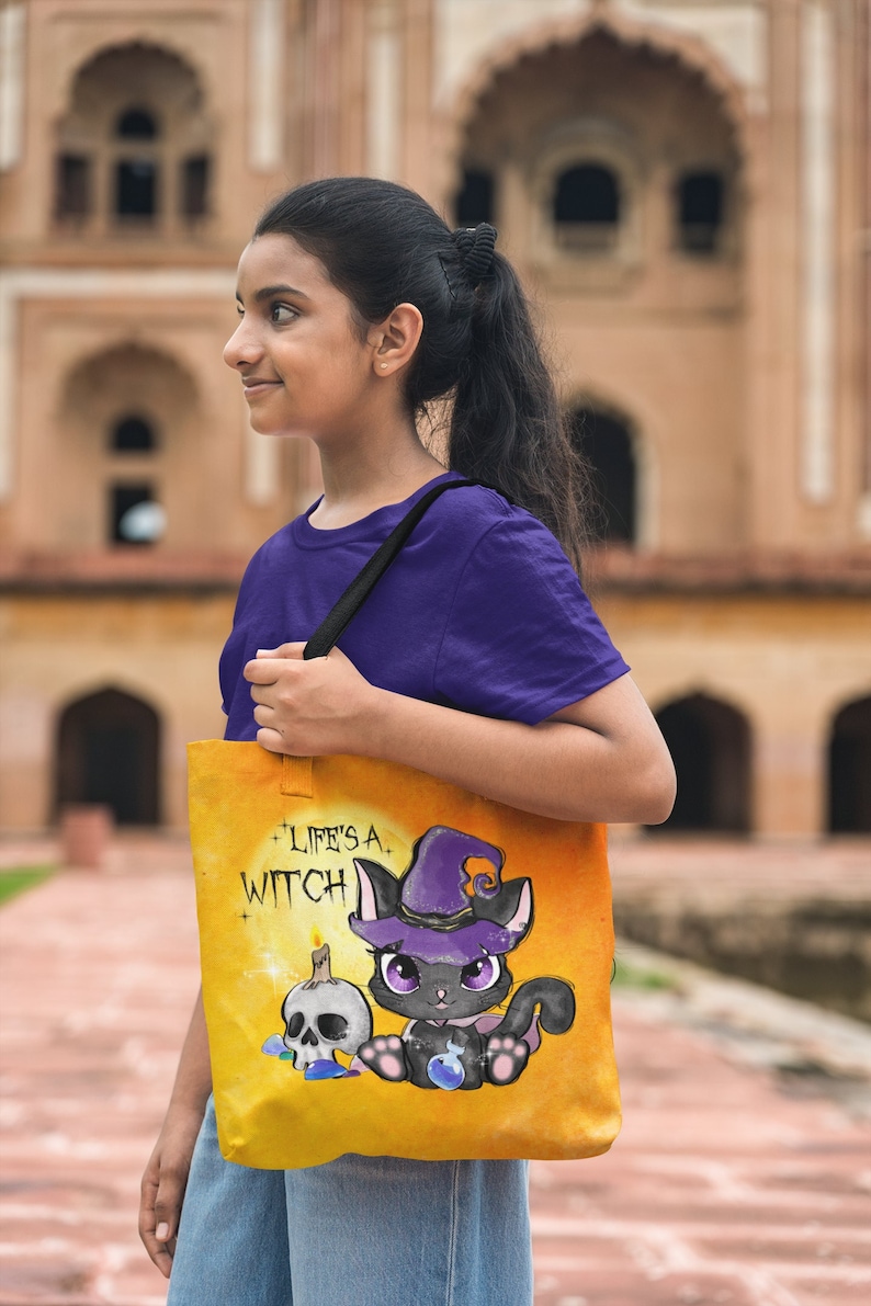 May include: A yellow tote bag with a black cat wearing a purple witch hat and holding a potion bottle. The bag has the text "Life's a Witch" printed on it.