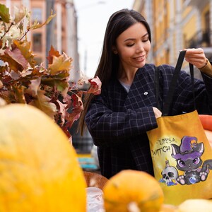 May include: A woman is holding a yellow tote bag with a black cat wearing a witch hat and the text "Life's a Witch". The bag is in front of a pile of pumpkins.