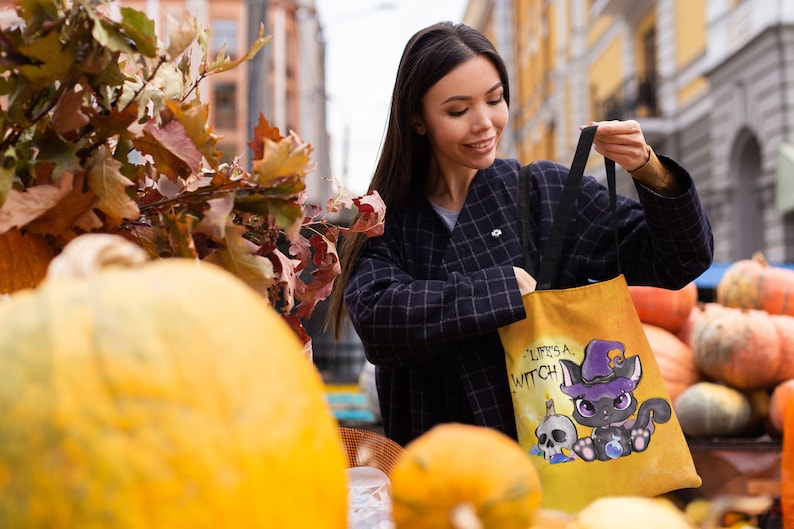 May include: A woman is holding a yellow tote bag with a black cat wearing a witch hat and the text "Life's a Witch". The bag is in front of a pile of pumpkins.