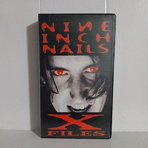 May include: A black VHS tape featuring the band Nine Inch Nails. The cover art displays a grayscale image of a person with red eyes and the text "NINE INCH NAILS" in red, with "X FILES" below.