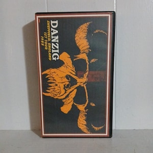 May include: A black cassette tape case featuring the band name "DANZIG" in white text. The case has an orange skull graphic and the text "Sheffield, England 1ST TOUR 1988".