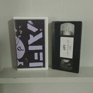 May include: A black VHS tape with the text "AFI Live Corona CA 1999" on the label. The tape is next to a black and white VHS tape with the text "AFI" on the label.