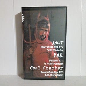 May include: A black VHS tape featuring a person in a devil costume holding a trident. The tape includes the band names SNOT, V.O.D., and Coal Chamber, along with venue and date information. The image is set against a white background.