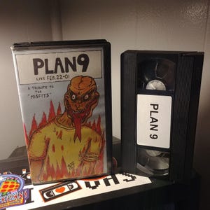 May include: A VHS tape with a black case and a label that says "PLAN 9" in white text. The tape is labeled with the text "PLAN 9 LIVE FEB. 22-01 A TRIBUTE TO THE "MISFITS"" and features a cartoon illustration of a monster with a long tongue.