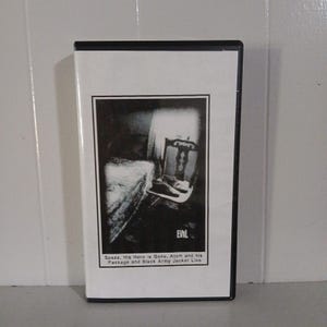 May include: A VHS tape with a black and white image of a chair and a bed. The text on the tape reads "EVIL Spazz. His Hero is Gone, Atom and his Package and Black Army Jacket Live."