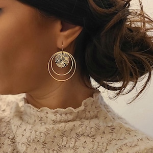 Könnte beinhalten: Goldene Creolen mit einem floralen Design auf dem Mittelstück. Die Ohrringe werden von einer Person mit langen braunen Haaren getragen.