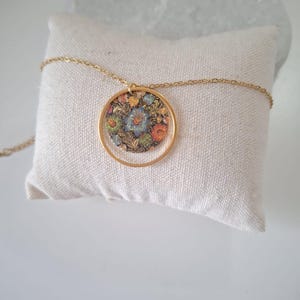 May include: A gold-colored necklace featuring a circular pendant with a floral design. The pendant has a gold frame and a colorful floral pattern with blue, orange, and green hues. The necklace is resting on a white pillow.