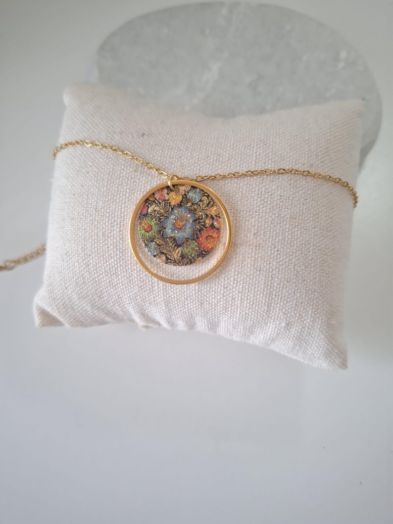 May include: A gold-colored necklace featuring a circular pendant with a floral design. The pendant has a gold frame and a colorful floral pattern with blue, orange, and green hues. The necklace is resting on a white pillow.