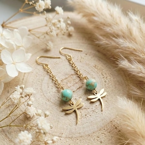 May include: A pair of gold-toned drop earrings featuring a delicate chain, a round turquoise-colored bead with dark veining, and a small gold-toned dragonfly charm at the base. The earrings are suspended from simple gold-toned ear wires.