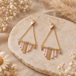 May include: A pair of gold-toned triangle-shaped drop earrings featuring a small white triangular accent at the top, delicate gold chains, and a horizontal bar adorned with small pale pink cylindrical beads and a central flat gold triangle charm.