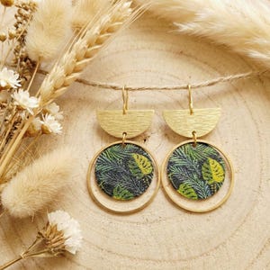Peut inclure: Une paire de boucles d'oreilles pendantes avec une pièce supérieure en demi-cercle doré brossé reliée à un grand anneau doré. À l'intérieur de chaque anneau se trouve un insert circulaire avec un motif de feuilles tropicales vert et jaune.