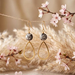 May include: A pair of drop earrings featuring a gold-toned hook, a round black bead with a white floral pattern, and a gold-toned teardrop-shaped frame. The earrings hang from a thin string suspended in front of light-colored dried foliage.