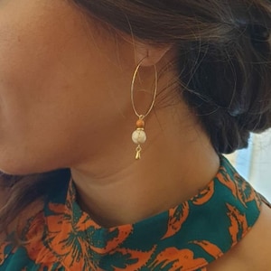 Peut inclure: Boucles d'oreilles créoles dorées avec un design pendant. Chaque boucle d'oreille comporte une pierre blanche et beige, une petite perle orange et un charm en forme de goutte d'eau dorée. La personne porte un haut à fleurs turquoise et orange.