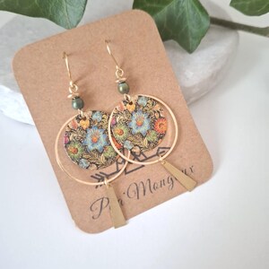May include: Gold-tone hoop earrings with floral patterned circular pendants and geometric brass accents. The earrings feature a small green bead near the hook. The earrings are displayed on a brown card with the text "P'i Mang ux".