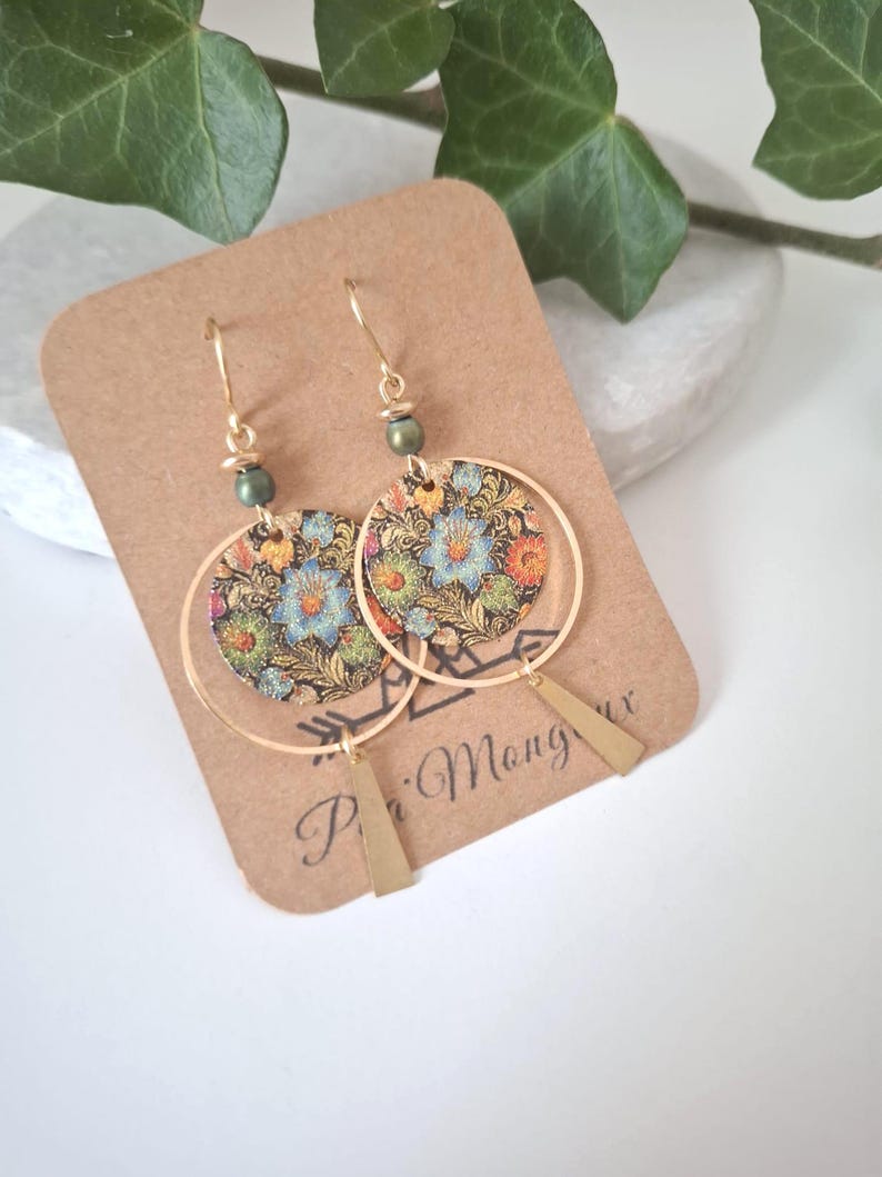 May include: Gold-tone hoop earrings with floral patterned circular pendants and geometric brass accents. The earrings feature a small green bead near the hook. The earrings are displayed on a brown card with the text "P'i Mang ux".