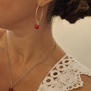 Peut inclure: Une boucle d'oreille créole en argent avec une petite perle rouge et un collier à chaîne en argent assorti avec un pendentif perle rouge. L'ensemble est porté avec un haut blanc doté d'une bordure en dentelle crochetée sur l'épaule.