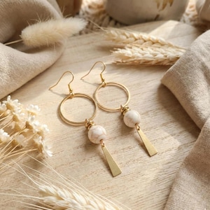 Peut inclure: Une paire de boucles d'oreilles pendantes dorées composées d'un anneau circulaire, d'une perle ronde à motif marbré blanc et d'une petite breloque triangulaire en métal doré suspendue à un crochet. Présentées sur une surface en bois clair.
