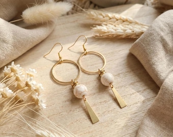 May include: A pair of gold-toned dangle earrings featuring a circular hoop, a round white marble-patterned bead, and a small triangular gold-toned metal charm suspended from a hook wire. The earrings are displayed on a light-colored wooden surface.