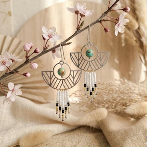 Peut inclure: Une paire de boucles d'oreilles pendantes argentées avec une monture métallique géométrique en demi-cercle, une perle ronde couleur turquoise et une frange de petites perles de verre blanches, noires et transparentes suspendues.