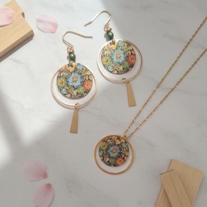 May include: A gold-toned jewelry set featuring floral patterned pendants and earrings. The set includes a necklace with a circular pendant, and a pair of earrings with a similar design. The floral pattern includes blue, orange, and yellow flowers on a dark background.