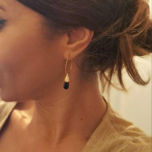 Peut inclure: Boucles d'oreilles créoles dorées avec une pierre noire en forme de goutte et un détail doré texturé. La personne porte les boucles d'oreilles avec des cheveux bruns attachés en chignon et un haut doré.