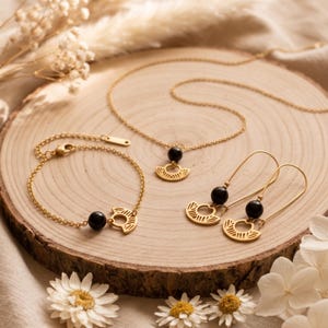 May include: A gold-toned jewelry set consisting of a necklace, a bracelet, and a pair of drop earrings. Each piece features a polished black spherical bead paired with a gold-toned semi-circular charm engraved with a sunburst line pattern.