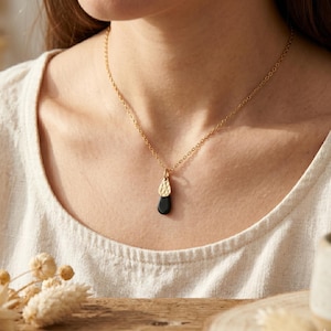 Peut inclure: Un collier doré avec un pendentif composé d'une partie supérieure dorée texturée et d'une partie inférieure noire en forme de goutte. Le collier est porté sur un haut crème.