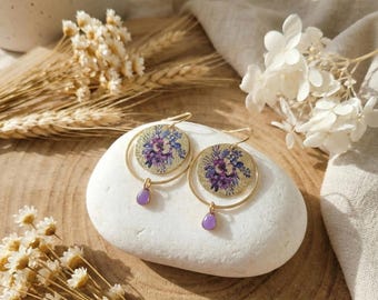 May include: A pair of gold-toned hoop earrings featuring a circular pendant with a purple and blue floral print. Each earring includes a small, dangling purple teardrop-shaped charm suspended from the bottom of the gold hoop frame.