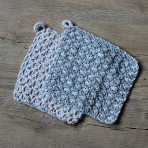 Puede incluir: Dos agarraderas de olla de ganchillo en azul y blanco. Las agarraderas están hechas con un patrón de punto sencillo y tienen un lazo para colgar.