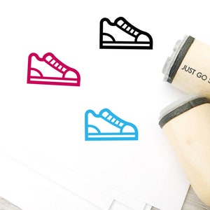 May include: Wooden stamps with the text "JUST GO STAMPS" and a sneaker icon in red, black, and blue. The stamps are on a light wood surface with white paper.