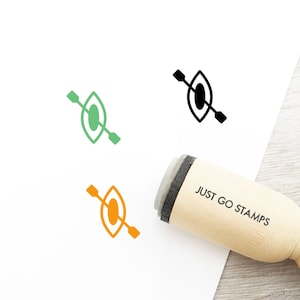 Op de afbeelding: Een houten rubberen stempel met de tekst "JUST GO STAMPS" en een zwart-witte illustratie van een kajak met een peddel. De stempel ligt op een wit oppervlak.