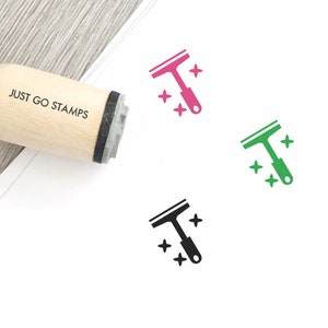 Op de afbeelding: Een houten stempel met de tekst "JUST GO STAMPS" erop. De stempel wordt gebruikt om een ontwerp van een rakel met glinsterende sterren te maken. Het ontwerp wordt in drie kleuren weergegeven: roze, groen en zwart.