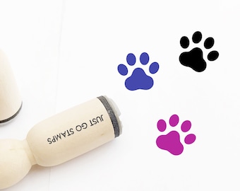 Puede incluir: Tres sellos de madera con diseños de huellas de patas. Los sellos son de color negro, morado y azul. Los sellos están etiquetados como "Just Go Stamps".