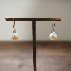 May include: A pair of drop earrings with large, lustrous, off-white pearls hanging from fine gold-coloured wires. The earrings are displayed on a gold-toned stand, highlighting their simple yet sophisticated design. The pearls have a soft, warm glow.