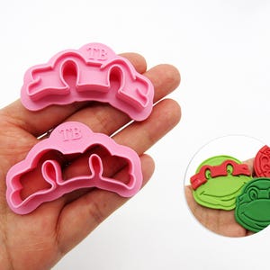 May include: Two pink plastic cookie cutters with the letters "TMB" are held in a hand. The cutters are in the shape of a cloud. A small image shows three colorful Teenage Mutant Ninja Turtle face-shaped cookie decorations.