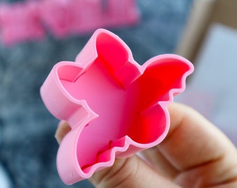 May include: Pink plastic cookie cutter shaped like a bat. The cutter is held in a hand.