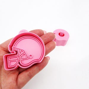 May include: Pink plastic cookie cutter shaped like a football helmet. The cutter has a raised design and is approximately 2 inches in diameter.